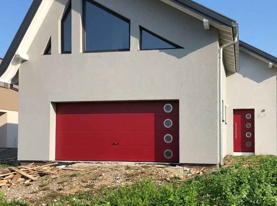 Façade de maison moderne en enduit gris clair, avec porte de garage sectionnelle rouge vif à rainures horizontales, équipée de 4 hublots ronds en aluminium brossé